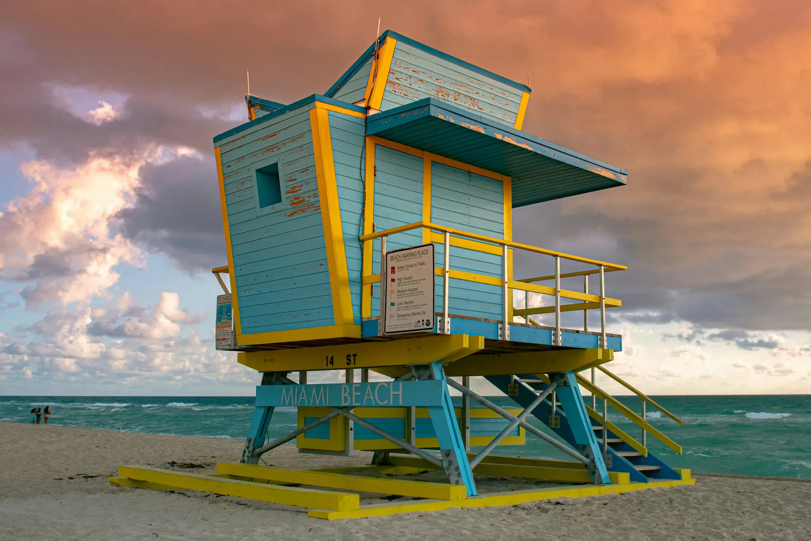 Kumsalda mavi-sarı boyalı art deco cankurtaran kulesi; üzerinde “MIAMI BEACH” yazıyor.