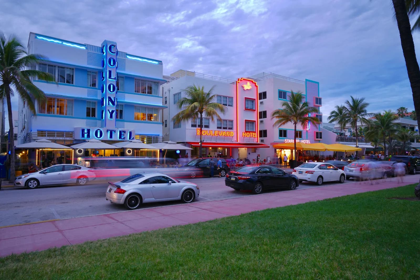 Ocean Drive’da mavi neon tabelalı Colony Hotel cephesi; önünden geçen arabalar ve kaldırım boyunca palmiyeler.